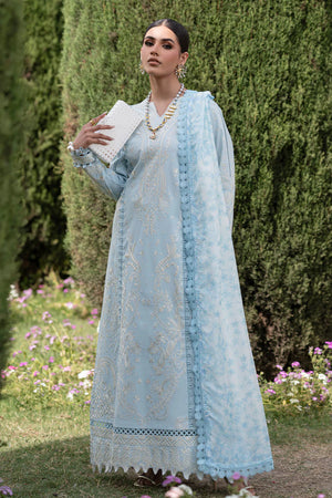 Woman in a light blue embroidered traditional outfit standing outdoors with greenery and flowers in the background