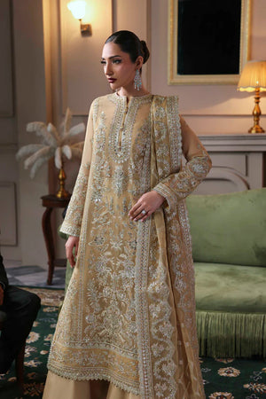 Woman in a traditional embroidered outfit standing in an elegant room with a green sofa and decorative elements. Tajmeen