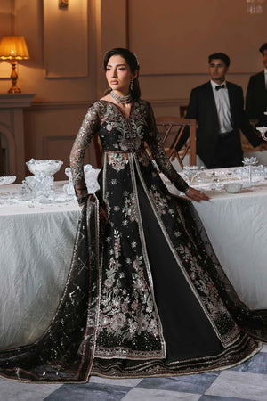 Woman in an elegant black embroidered gown standing in a formal setting with table settings and people in the background. Zarafreen